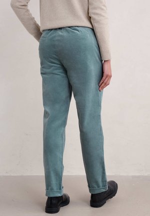 Persona con pantalones de pana en color verde azulado arremangados en los puños, suéter beige y botas negras, de pie contra una pared clara y sencilla.