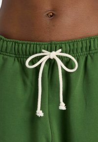 Grüne Baumwoll-Jogginghosen mit einem elastischen Bund und einem weißen Kordelzug, der zu einer Schleife gebunden ist. Glatte Textur mit gerafften Stoffdetails.