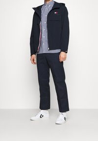 Giacca impermeabile blu navy con cappuccio, abbinata a una camicia a quadri blu e bianca, pantaloni scuri e sneakers bianche con dettagli neri.