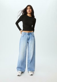 Schwarzes Langarmoberteil kombiniert mit hellblauen, weiten Jeans, ausgestattet mit einem silbernen Knopf und Taschen, stilvoll ergänzt mit weißen Sneakers.