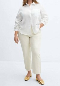 Mujer vestida con una camisa blanca de botones, pantalones rectos color crema con dobladillos deshilachados y bailarina dorada con pequeños tacones.