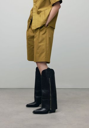 Bottes en cuir noires, hautes, avec un bout pointu et un motif gaufré ; dotées d'une fermeture éclair sur le côté et d'un talon épais. Associées à une tenue moutarde.
