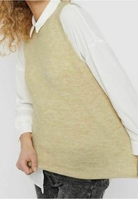 Maglione smanicato in beige chiaro lavorato a maglia con una consistenza morbida, stratificato su una camicia bianca con colletto, abbinato a jeans scuri.
