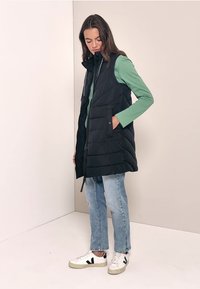 Gilet imbottito nero con chiusura a zip, tasche laterali e colletto alto, indossato sopra una camicia verde a maniche lunghe e abbinato a jeans chiari e sneaker.