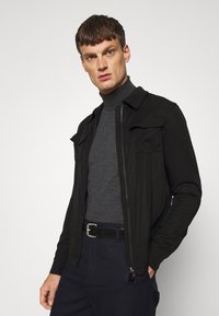 Veste noire zippée avec deux poches avant, portée par-dessus un col roulé gris foncé, complétée par une ceinture et un pantalon sombre. Texture de tissu lisse.