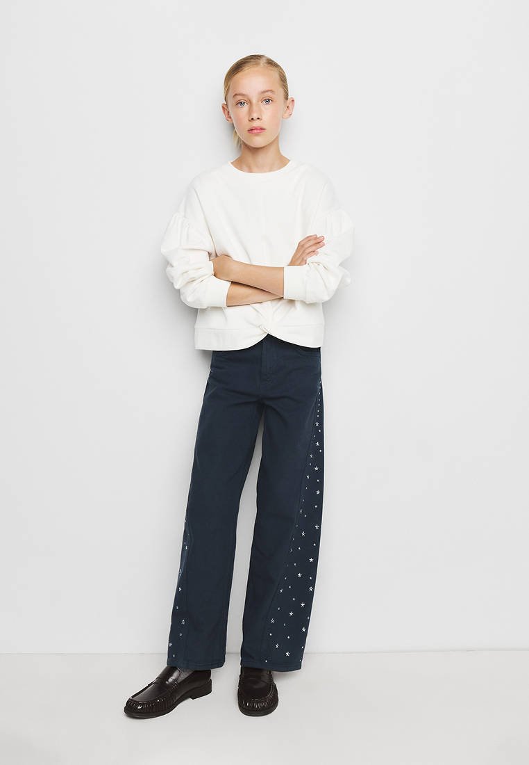 Jeune personne debout, les bras croisés, portant un haut blanc à manches longues, un pantalon bleu foncé avec des motifs d'étoiles sur les côtés et des chaussures noires.