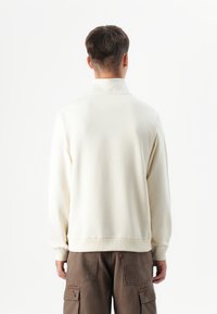 Ung person med kort hår, som vender væk, iført en lys kremfarvet sweatshirt og brune cargo-bukser mod en simpel hvid baggrund.