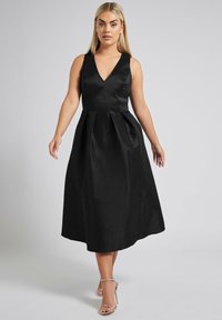 Musta satiini mekko, jossa on syvä V-pääntie, hihaton malli, laskostettu hame ja polven alapuolelle ulottuva pituus. Käytetään hopeisten remmikenkien kanssa.