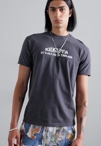 Gray cotton t-shirt with white text "KIDSUPER IT'S ALL UP IN THE AIR" on the front. Model also wears patterned shorts. Smooth texture.