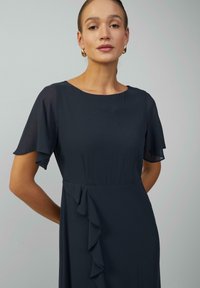 Marinablauwe jurk met korte, doorschijnende mouwen. Heeft een subtiele ruchedetail aan de zijkant, gemaakt van lichte stof met een gladde textuur.