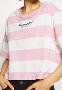 Růžové a bílé pruhované zkrácené tričko, s kulatým výstřihem a vyšitým logem "Superdry" na přední straně. Měkký bavlněný materiál.