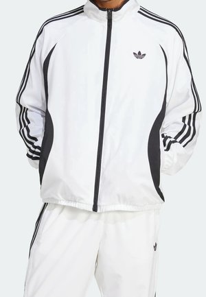 Osoba ubrana w biały dres Adidas z czarnymi paskami, składający się z bluzy na suwak i dopasowanych spodni z logo na klatce piersiowej i udzie.