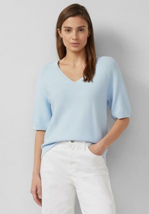 Kobieta o brązowych włosach, stojąca na tle szarej ściany, ubrana w jasnoniebieski sweter z krótkim rękawem i dekoltem w serek oraz białe spodnie, z jedną ręką w kieszeni.