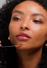 Femme aux cheveux bouclés appliquant un fond de teint liquide avec un pinceau sur son menton et ses joues, montrant le processus d'application du maquillage.