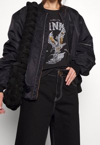 Černá bombojaketa s ribovanými manžetami, nošená přes černé a stříbrné tričko s potiskem. Černé kalhoty s širokými nohavicemi a kontrastním šitím, držící texturovanou černou kabelku.