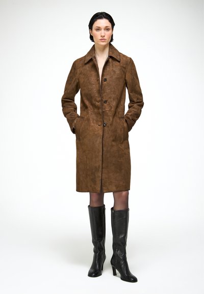 Femme debout portant un long manteau en daim marron avec des boutons et des bottes noires à talons montant jusqu'aux genoux, les mains dans les poches du manteau.
