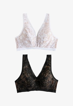 Donna che indossa un reggiseno in pizzo nero e biancheria intima coordinata in pizzo, in piedi su uno sfondo chiaro con le mani sui fianchi.