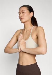 Sutiã desportivo bege com alças finas, textura suave e detalhes em metal circulares. Modelo posa com as mãos juntas, destacando um design minimalista.