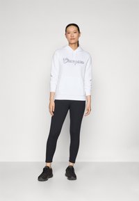 Felpa bianca con cappuccio e logo "Champion", abbinata a leggings neri e scarpe da ginnastica nere, modello in piedi contro uno sfondo neutro.