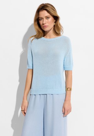 Kobieta ubrana w jasnoniebieski dzianinowy sweter z krótkim rękawem oraz dopasowane szerokie spodnie, uzupełnione złotą bransoletką i pierścionkiem.