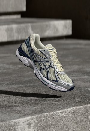 Zapato de correr ligero con parte superior de malla en color crema y marino, cuenta con un cuello acolchado, superposiciones texturizadas y una suela amortiguada.