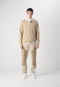 Un hanorac bej cu guler alb și logo, asortat cu pantaloni bej deschis cu pet-uri din catifea la față. Adidași albi completează ținuta.