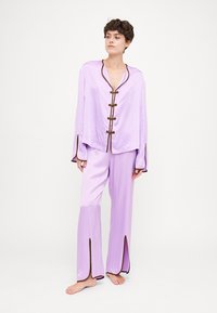 Ensemble de pyjama en satin lilas comprenant un haut à manches longues avec un décolleté arrondi, des fermetures à boutons décoratifs marron, et un pantalon à jambes larges avec des fentes latérales.