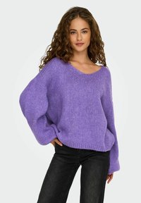Maglione viola a maglia con scollatura ampia, maniche lunghe e vestibilità comoda. Presenta un tessuto morbido e testurizzato. Indossato con jeans neri.