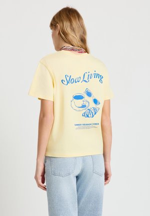 Donna con capelli lunghi che indossa una maglietta gialla con il testo "Slow Living" e illustrazioni blu di caffè, occhiali da sole e croissant sul retro.
