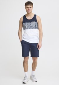 Námornícky tank top s bielymi detailmi a texturovaným vzorom na hornej časti, v kombinácii s tmavomodrými šortkami. Model má na sebe sivé tenisky a biele členkové ponožky.