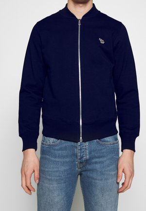 Mann trägt eine marineblaue Bomberjacke mit Reißverschluss und kleinem Zebra-Emblem auf der Brust sowie hellblaue Jeans, steht vor einem einfarbigen Hintergrund.