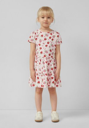 Młoda dziewczyna z blond włosami w krótkim różowym sukienka z czerwonym nadrukiem truskawek i białych trampkach, stoi na tle jasnoszarym.