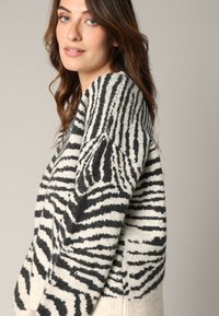 Pull en tricot à motifs zébrés noir et crème, avec un col en V et des manches longues, offrant une coupe décontractée. Texture douce et coutures visibles.