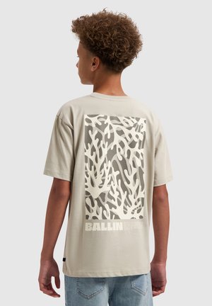 Tienerjongen met krullend haar, die een beige T-shirt draagt met witte koraalachtige patronen en de tekst "BALLIN" op de rug, gecombineerd met een blauwe spijkerbroek.