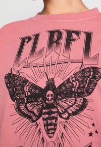 Camiseta de algodón rosa con un gran gráfico negro de una polilla calavera, con alas detalladas y rayos detrás, complementada con texto en negrita.