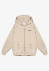 Beige zip-up hoodie tillverkad av mjukt tyg. Har huva, framfickor, ribbade muddar och en svart logotyp på vänstra bröstområdet.