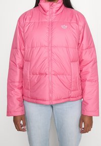 Rosa puffjacka med hög krage, horisontell quiltad design och en liten vit logotyp på vänster bröst. Har sidofickor.