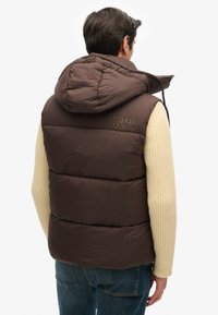 Braune wattierte Weste mit Kapuze, die ein gestepptes Muster und das "SUPERDRY"-Logo trägt. Ergänzt durch einen cremefarbenen Strickärmel und dunkle Jeans.