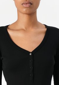 Mustapaitainen bodysuit, jossa on V-mallinen kaula-aukko ja edessä neljä nappia. Kangas vaikuttaa sileältä ja istuu tiukasti.