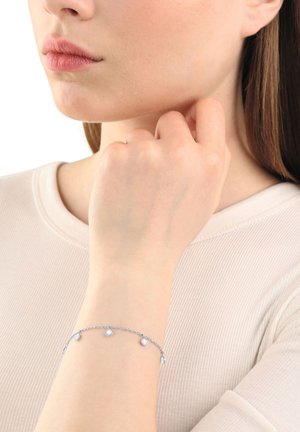 Pulsera de cadena de plata con cuentas redondas y transparentes espaciadas uniformemente a lo largo de la longitud, llevada en una mano sobre un fondo de tela de color claro.