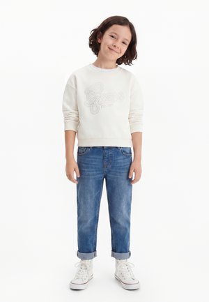 Bambino con capelli scuri fino alle spalle sorride, indossa una felpa bianca Guess, jeans blu risvoltati e sneakers bianche alte.