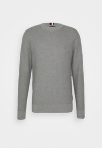Pull gris en tricot avec manches longues, encolure ronde et poignets côtelés. Présente un petit logo sur la poitrine et un motif texturé.
