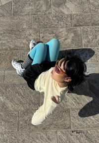 Persona oranžos saulesbrillēs, gaiši dzeltenā garroku kreklā, zilās legingos, svītrainās zeķēs un baltās sporta kurpēs sēž uz akmens bruģa.