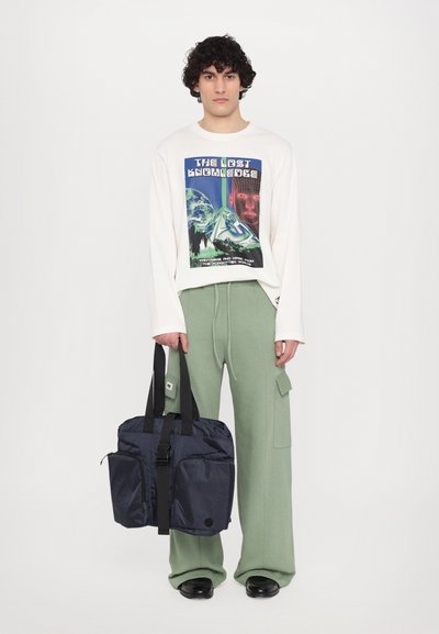 Navy fabric bag with two front pockets, black straps, worn by a model in an oversized white graphic long-sleeve shirt and green pants.
