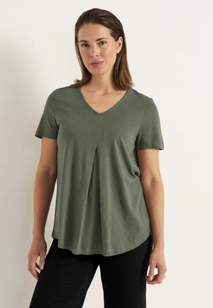 Frau trägt ein olivgrünes V-Ausschnitt-Kurzarmshirt mit Frontfalte, kombiniert mit schwarzen Hosen, steht vor hellem einfarbigem Hintergrund