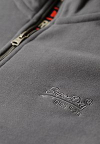 Sudadera gris con cremallera fabricada en tejido suave, con un logotipo bordado destacado en el pecho y una cremallera de metal. Acabado suave y texturizado.