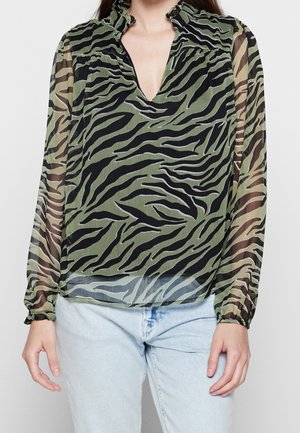 Vrouw die een doorschijnende blouse met lange mouwen draagt, met een groene en zwarte zebraprint en een geraffelde kraag, gecombineerd met lichtblauwe jeans.