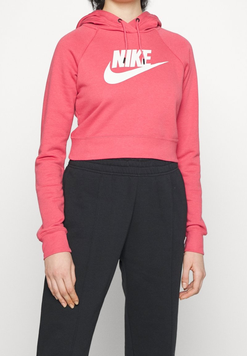 Růžová krátká mikina s bílým logem Nike na hrudi, s kapucí na stahovací šňůrku a dlouhými raglánovými rukávy. Doplněná tmavými tepláky.