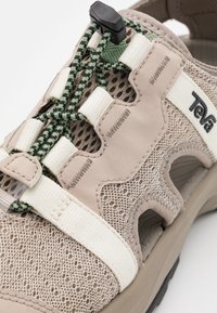 Světle béžová bota z síťovaného materiálu s pevnými popruhy. Obsahuje gumovou podrážku, zelené a černé tkaničky a výrazné logo na boku.