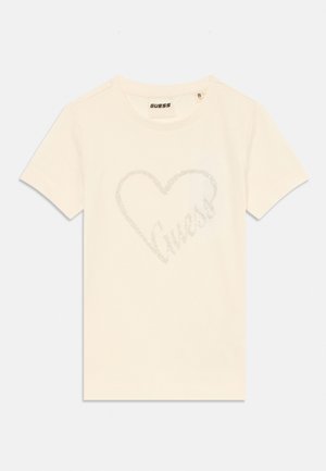T-shirt de algodão cor creme com um design de coração cintilante contendo a palavra "Guess" no interior, mangas curtas e gola redonda.